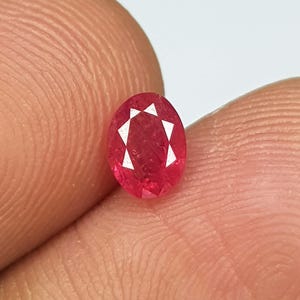 Peut inclure: Une pierre précieuse de type rubis, taillée en forme ovale. Le rubis est d'une couleur rouge vif avec des facettes internes blanches. La gemme est tenue entre deux doigts, mettant en valeur sa taille et sa clarté. La forme du rubis est allongée, avec une surface polie.