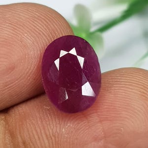 Puede incluir: Una piedra preciosa de rubí de forma ovalada y tallada con facetas, de un color rojo magenta profundo y saturado. La piedra presenta un corte claro y simétrico con múltiples facetas triangulares que reflejan la luz.
