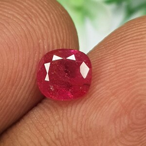 Peut inclure: Une pierre précieuse rubis taille coussin, d'une teinte rouge profond avec une surface à facettes. La pierre présente des caractéristiques de clarté interne et des facettes réfléchissant la lumière, offrant une finition polie et une couleur rouge intense.
