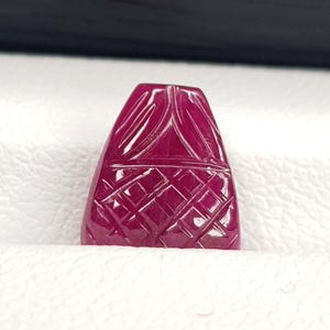 May include: A carved, deep red gemstone with a geometric pattern. The gemstone has a triangular shape with a flat base and is set against a white background. The surface of the gemstone is highly polished.