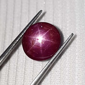 May include: A round, polished ruby gemstone, held by tweezers. The gemstone is a deep red color with a six-rayed star effect. The tweezers are silver and the background is a light, textured surface.