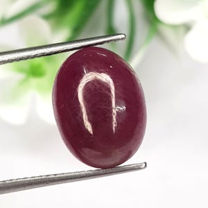 May include: A deep red, oval-shaped cabochon gemstone held by silver-toned metal tweezers. The stone features a smooth, polished surface with a visible internal structure and a distinct light reflection across its rounded top, showcasing a rich, saturated colour.