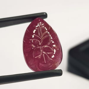 May include: A teardrop-shaped, deep red gemstone with a carved floral design. The ruby is held by black tweezers against a white background. The gemstone's surface reflects light, highlighting the intricate details of the carving.