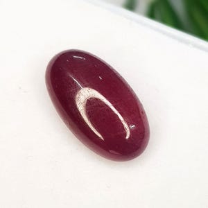 Könnte beinhalten: Ein tiefroter, oval geformter Edelstein mit einer glatten, polierten Cabochon-Oberfläche. Der Stein zeigt einen satten, gesättigten Burgunderfarbton und weist eine helle weiße Lichtreflexion auf der gewölbten Oberseite auf.