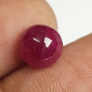 Op de afbeelding: Een diep magentarode edelsteen met een gladde, ronde cabochon-slijpvorm. De steen heeft een gepolijst, reflecterend oppervlak met subtiele interne insluitingen en een doorschijnende kwaliteit die het licht opvangt en een rijke kleur toont.