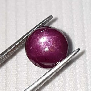 May include: A deep red, polished gemstone, held by silver tweezers. The gem has a star-like effect on its surface. The background is a white, textured surface. The gem is a cabochon cut.