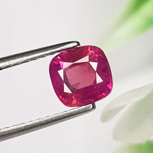 Op de afbeelding: Een edelsteen in kussenslijpvorm met een levendige, dieproze-rode kleur en een gefacetteerd oppervlak. De steen wordt vastgehouden door een zilverkleurige metalen pincet, wat de helderheid en geometrische vorm benadrukt tegen een wazige achtergrond.