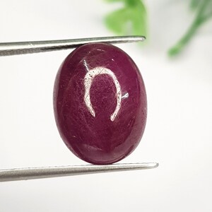 Könnte beinhalten: Ein ovaler, tief magentaroter Edelstein mit einer glatten, polierten Cabochon-Oberfläche. Der Stein weist in der Mitte eine deutliche, hell gefärbte, U-förmige Einschließung oder Reflexion auf und wird von einer silberfarbenen Metallpinzette gehalten.