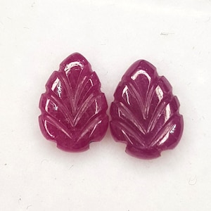 Op de afbeelding: Twee diep magenta, peervormige edelstenen gesneden met een symmetrisch bladachtig patroon. Elke steen heeft een gepolijst, gefacetteerd oppervlak met duidelijke ribbels die een visgraatontwerp creëren dat licht reflecteert.
