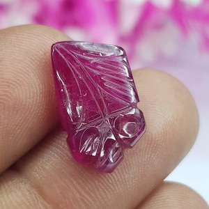 May include: A carved, deep red gemstone with intricate leaf-like patterns. The polished surface reflects light, highlighting the detailed carvings. The gemstone is held in a hand, showcasing its size and shape.