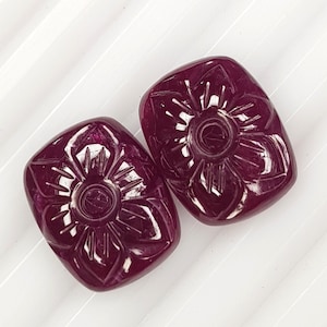 May include: Two deep magenta, rectangular-shaped gemstones with rounded corners, each featuring a carved, symmetrical floral pattern with radiating petals and a central circular indentation, displaying a polished, reflective surface texture.