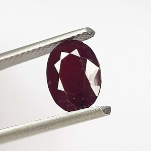 May include: A faceted, oval-shaped ruby gemstone held by silver tweezers. The ruby is a deep, rich red color with bright white facets. The gemstone is displayed against a plain white background.