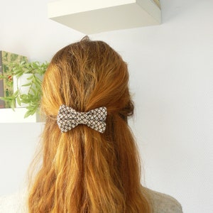 May include: A brown and white patterned bow hair clip is secured in the middle of long, wavy, auburn hair. The hair clip is a small bow shape. The person is wearing a gray and white striped sweater.