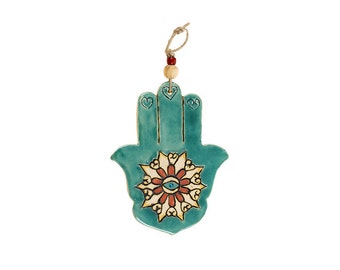 May include: Teal blue ceramic Hamsa hand with a white and orange mandala design featuring a blue evil eye in the center. The Hamsa is hanging from a string with a wooden bead.