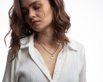 May include: A woman with brown wavy hair wearing a white collared shirt with a silver chain necklace with a circular pendant.