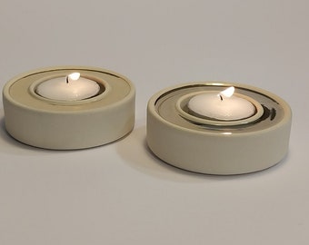 May include: Two white ceramic candle holders with a silver inner ring. Each holder has a small white tea light candle burning inside.