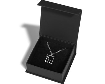 May include: A silver-colored pendant necklace with a geometric design, presented in an open black gift box with a soft, dark interior. The necklace features a square pendant with an abstract shape, suspended from a delicate chain.