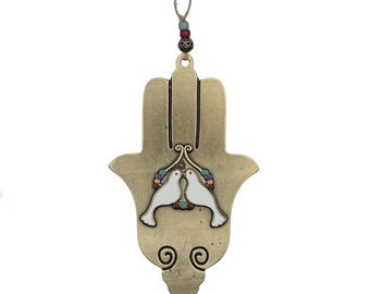 May include: A gold-colored Hamsa hand-shaped wall hanging with a decorative design. Two white doves are centered on the hand, surrounded by colorful beads. The hanging has a jute string for display.