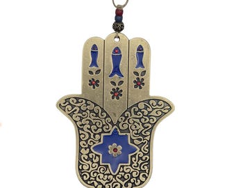 May include: A bronze-colored Hamsa hand-shaped wall hanging with intricate scrollwork. The hand features three blue fish and red flower accents. A blue star-shaped design is centered on the palm. It is suspended by a string with beads.