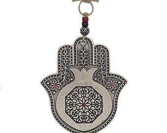 May include: A silver-colored Hamsa hand-shaped wall hanging with a pomegranate design. The hand features intricate black detailing and red accents. It is suspended by a natural fiber cord with decorative beads.