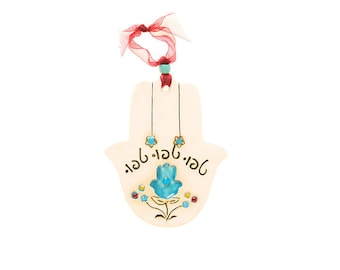 May include: Cream-colored Hamsa hand-shaped wall hanging with a turquoise blue hand design and floral accents. The Hamsa has a red ribbon for hanging and decorative text. The design includes small blue and gold star-shaped accents.