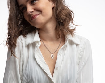 May include: A woman wearing a white collared shirt with a silver chain necklace and a geometric pendant.