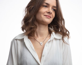 May include: A woman wearing a white collared shirt with a silver heart necklace featuring a cross design.