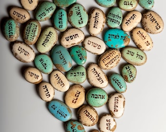 May include: A heart-shaped arrangement of small, oval stones. Each stone is a mix of colors, including white, beige, and turquoise, with black Hebrew text. The stones are arranged to form a heart shape, with various words inscribed on them.