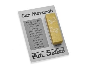 May include: A gold-colored mezuzah with a white inscription in Hebrew. The mezuzah is on a white card with the text "Car Mezuzah" and "Adi Sidler" in black.