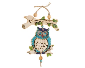 May include: A ceramic owl with blue and white paint, perched on a branch with green leaves. The owl has large, round eyes and a small beak. The branch is attached to a string with wooden beads.