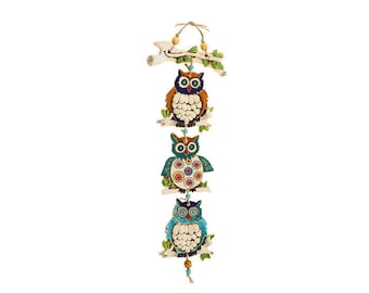 May include: A decorative hanging featuring three owl figurines in shades of blue, brown, and green. Each owl is perched on a branch, with a wooden accent at the top and a beaded tassel at the bottom. The owls have detailed patterns.