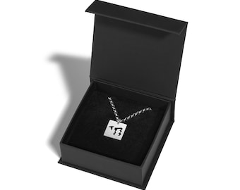 May include: A silver-colored pendant necklace with a square pendant featuring a stylized design, presented in an open black gift box with a black interior. The necklace chain is also silver. The box is on a white background.