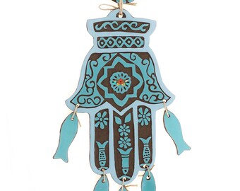 May include: A blue ceramic hamsa hand with brown details and a small blue flower design. The hamsa hand has five dangling blue fish charms and a small blue circle with a brown center at the top.