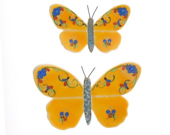 May include: Two yellow ceramic butterflies with blue and red floral designs. The butterflies are painted with intricate details and have a textured finish.