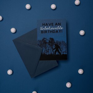 Peut inclure: Une carte bleue avec une enveloppe blanche. La carte pr&eacute;sente une silhouette noire de Bigfoot marchant dans une for&ecirc;t avec le texte "Have an Unbelievable Birthday!" en blanc.