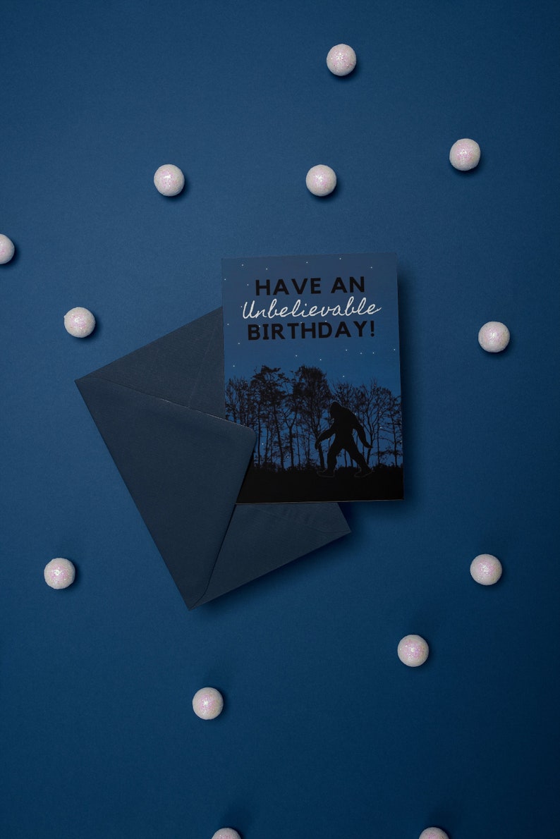 Peut inclure: Une carte bleue avec une enveloppe blanche. La carte pr&eacute;sente une silhouette noire de Bigfoot marchant dans une for&ecirc;t avec le texte "Have an Unbelievable Birthday!" en blanc.