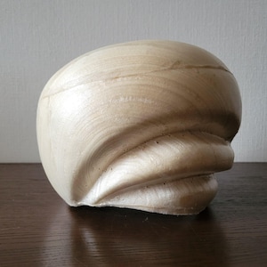 May include: A light-colored, wooden sculpture with a smooth, curved design. The wood grain is visible, and the piece has a rounded top and a flowing, wave-like form on the lower half. The sculpture is displayed on a dark surface.