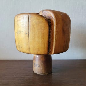 May include: A vintage wooden hat block with a square top and a rounded base. The block is made of light brown wood with visible grain and some dark brown markings. The base has the number "B-54" written on it.