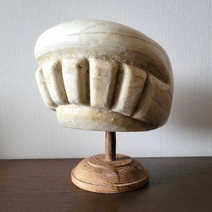 May include: A carved stone sculpture of a stylized mouth, mounted on a wooden stand. The sculpture is off-white with brown veining and a textured surface. The base is a round, light-colored wood.