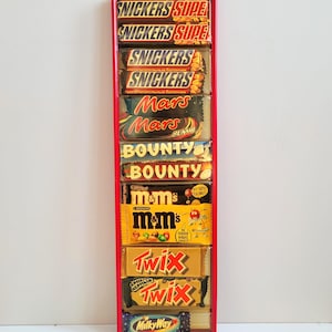 Könnte beinhalten: Eine rot gerahmte Präsentation mit verschiedenen Schokoriegeln, darunter Snickers, Mars, Bounty, M&M's, Twix und Milky Way. Die Riegel sind vertikal angeordnet und zeigen ihre bunten Verpackungen und Markennamen.