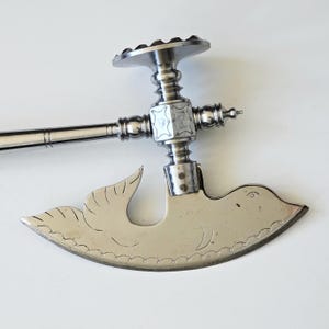 Op de afbeelding: Een zilverkleurig, antiek kaasmes met een vogelvormig lemmet. De handgreep heeft sierlijke details en een kruisvormig ontwerp. Het lemmet heeft een geschulpte rand en geëtste vogeldetails. Een uniek keukengerei.