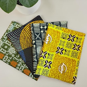 Op de afbeelding: Een verzameling kleurrijke Afrikaanse waxprintstoffen. De stoffen hebben verschillende patronen, waaronder geometrische vormen, bloemmotieven en traditionele symbolen. Het kleurenpalet omvat tinten groen, geel, zwart en oranje.