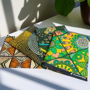 Op de afbeelding: Een verzameling kleurrijke, bedrukte stoffen in een waaier. De stoffen hebben geometrische en bloemmotieven in oranje, geel, groen en blauw. Op één stuk staat het woord "GUARANTEED" gedrukt.