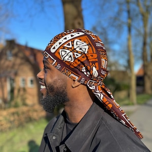 Op de afbeelding: Een persoon draagt een hoofddoek met een bruin en oranje patroon met witte geometrische ontwerpen. De doek is aan de achterkant van het hoofd in een knoop geknoopt en de uiteinden hangen naar beneden.