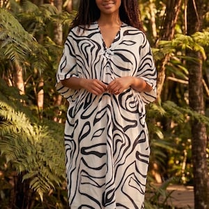 May include: A flowing, V-neck dress with a black and white abstract pattern. The dress has short sleeves and a decorative clasp at the neckline. The model is standing in a lush, green outdoor setting.