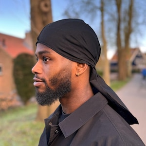 Op de afbeelding: Een man draagt een zwarte durag en een zwart jack. De durag is vastgebonden aan de achterkant van zijn hoofd en hangt naar beneden over zijn rug.