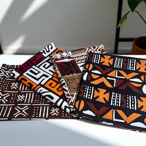 Op de afbeelding: Een verzameling gevouwen stoffen met gedurfde, geometrische patronen in bruin, oranje, wit en bordeaux. De stoffen hebben verschillende ontwerpen, geschikt voor woondecoratie of knutselen.