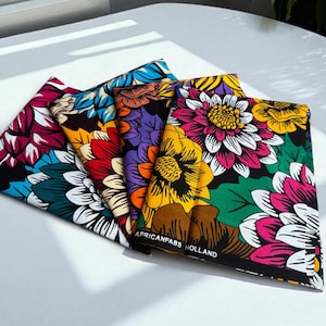 Op de afbeelding: Een stapel kleurrijke stoffen met bloemenpatronen. De stoffen tonen grote bloempatronen in roze, geel, blauw, rood en paars. De tekst "AFRICANFABS HOLLAND" is zichtbaar op een van de stoffen.