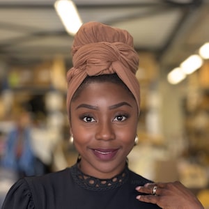 Pode incluir: Uma mulher com pele marrom escura está usando um turbante marrom. Ela está sorrindo e olhando diretamente para a câmera. Ela está vestindo uma camisa preta com uma renda ao redor do pescoço.