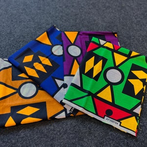 Op de afbeelding: Vier vierkante stukken stof met geometrische patronen in zwart, geel, rood, groen, blauw en wit. De stofvierkanten zijn gevouwen en in een waaier vorm gerangschikt. De stof heeft de tekst "AF200704" erop gedrukt.